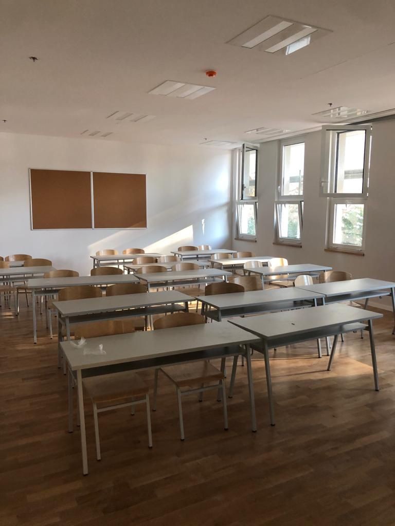 Empty classroom with wooden flooring, several desks, chairs, and a bulletin board on the wall.